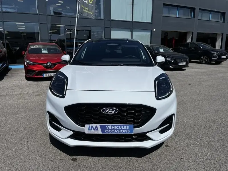 Vue frontale d'un Ford Puma blanc 2025 avec calandre noire et badge Ford, stationné devant un bâtiment commercial.