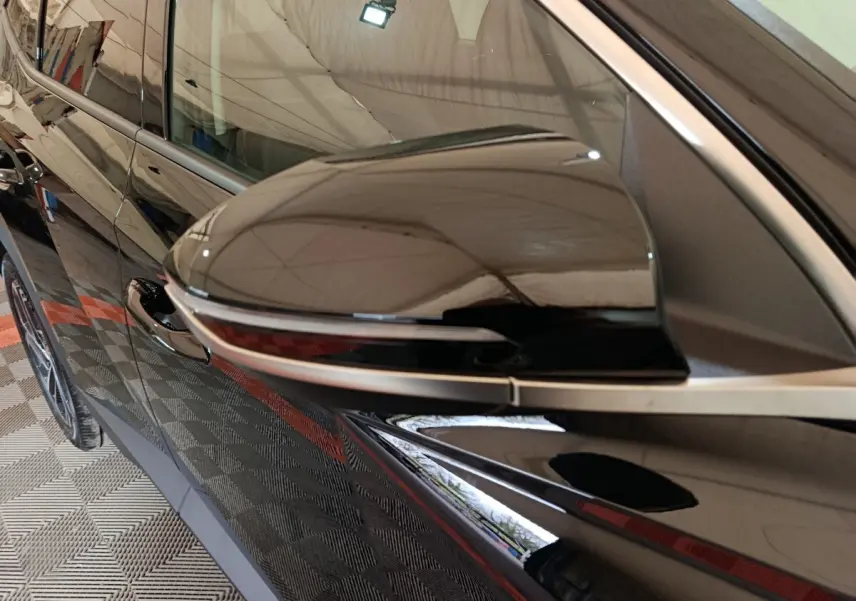 Gros plan sur le rétroviseur côté gauche noir brillant du Hyundai Tucson 1.6 Hybrid 2025, avec reflet de l’environnement.