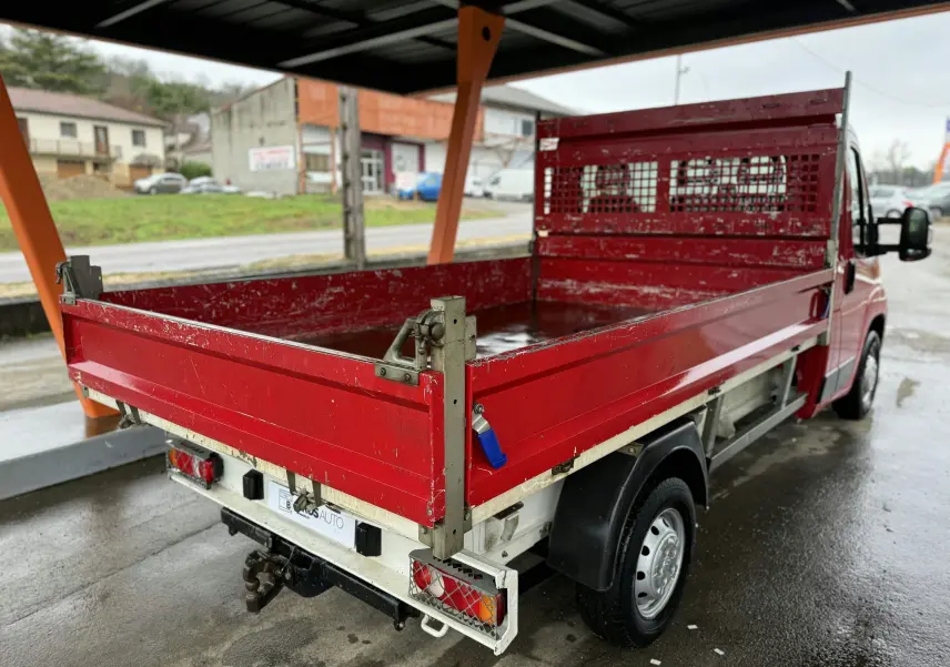 Vue 3/4 arrière droite d'un Citroën Jumper benne rouge avec ridelles métalliques et attelage sous abri.