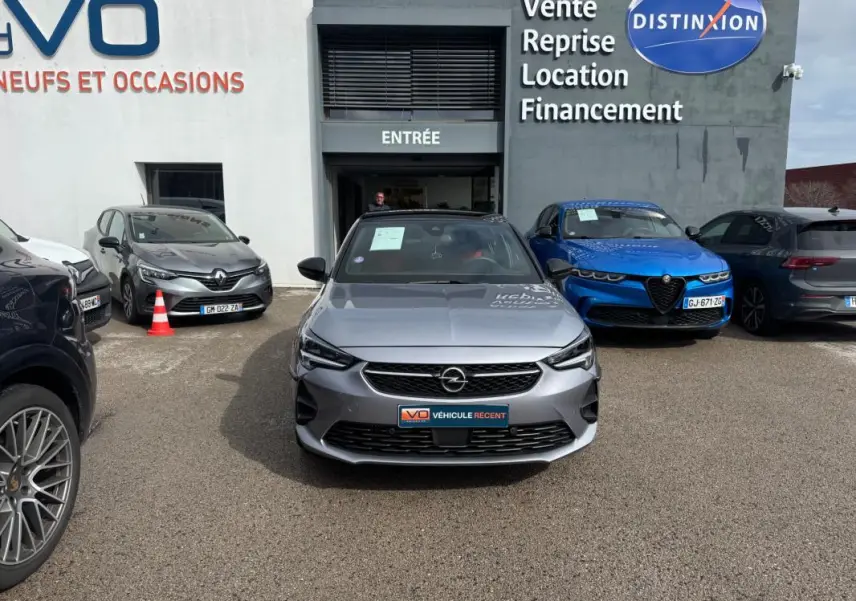 Vue avant d'une Opel Corsa gris clair 2022 garée devant un concessionnaire avec logo et panneaux visibles.
