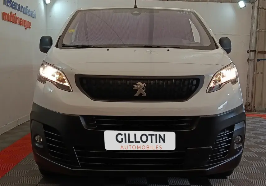 Vue de face d'un Peugeot Expert Fourgon blanc avec calandre noire et phares allumés dans un garage.