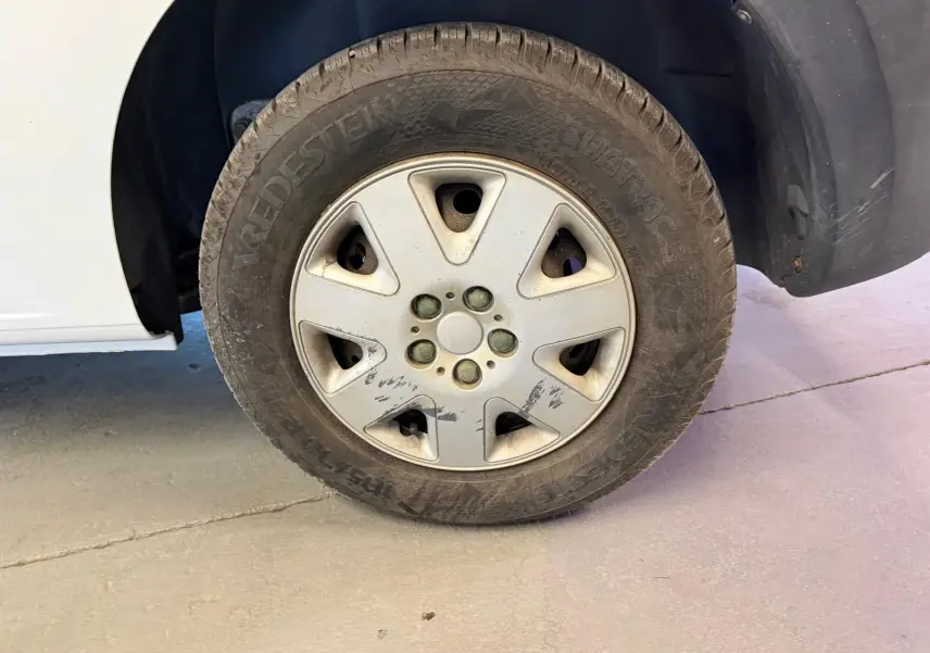 Gros plan sur la roue avant droite d’un Renault Kangoo blanc de 2011, jante avec traces d’usure visibles.