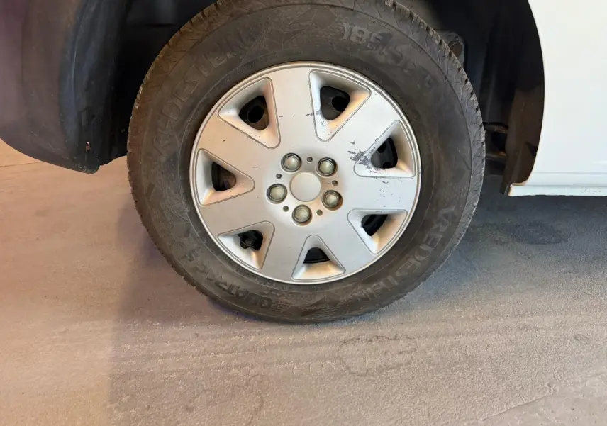 Gros plan sur la roue avant droite d’un Renault Kangoo blanc de 2011, montrant la jante en alliage avec quelques rayures.