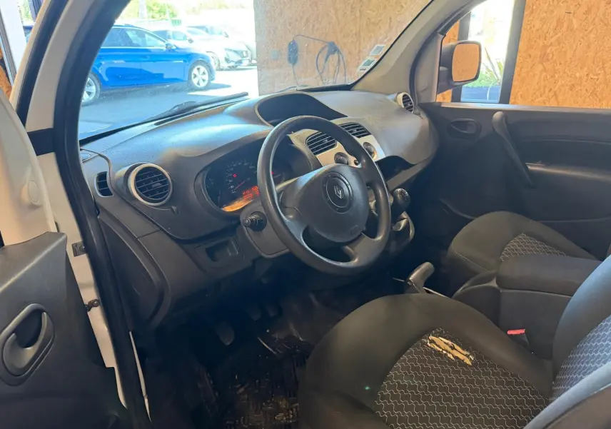 Vue intérieure côté conducteur du Renault Kangoo 1.5 DCI blanc, montrant le tableau de bord et les sièges en tissu noir.