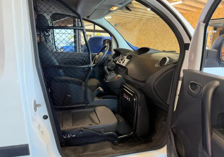 Vue intérieure côté conducteur du Renault Kangoo blanc 2011 avec cloison métallique séparant l'habitacle et siège en tissu noir.