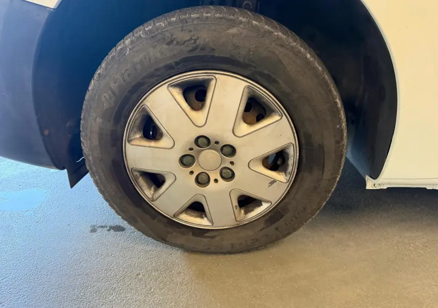Gros plan sur la roue avant gauche d'un Renault Kangoo blanc de 2011, avec jante en alliage et pneu usé.