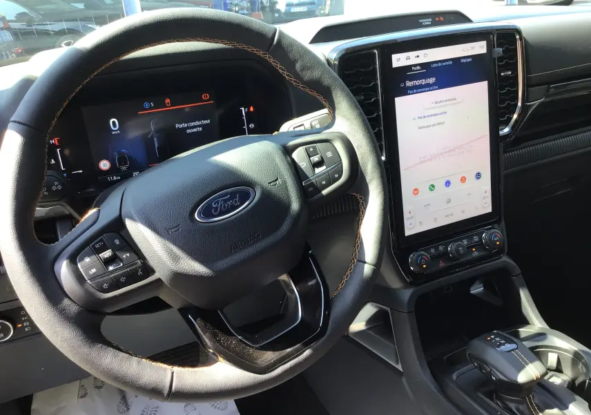 Vue intérieure du Ford Ranger Wildtrak gris carbone, mettant en valeur le volant cuir avec surpiqûres orange et l'écran tactile vertical.