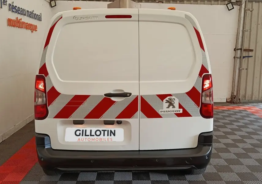 Vue arrière d’un Peugeot Partner blanc avec bandes réfléchissantes rouges et blanches et logo visible sur porte arrière.