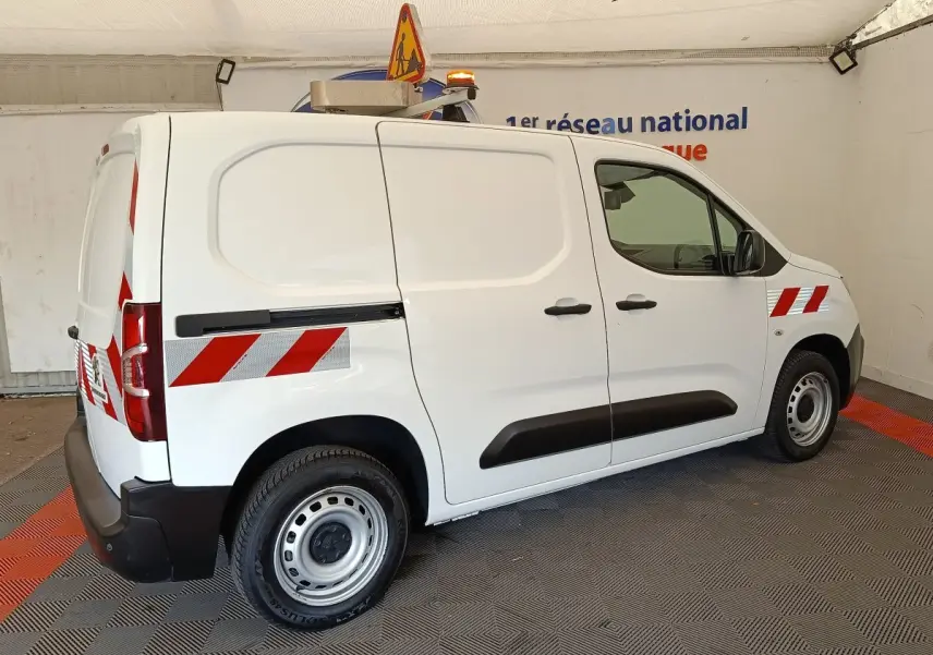 Vue 3/4 arrière droite d’un Peugeot Partner blanc 2022 avec bandes réfléchissantes rouges et gyrophare orange.