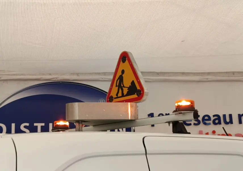 Vue partielle du toit blanc du Peugeot Partner 2022 avec panneau triangulaire de chantier et balises lumineuses orange allumées.