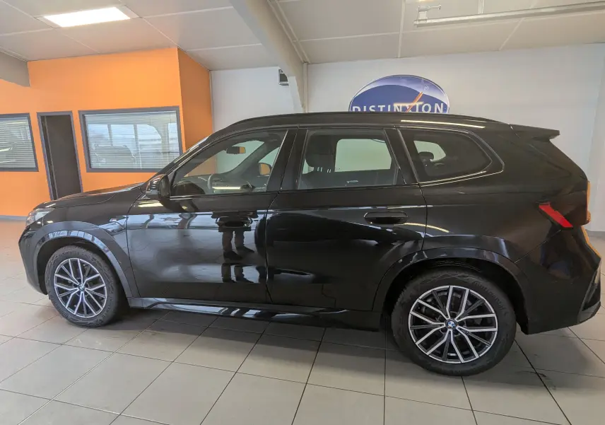 BMW X1 noir vue de profil côté gauche, avec jantes alliage bicolores et barres de toit individuelles visibles en intérieur showroom.