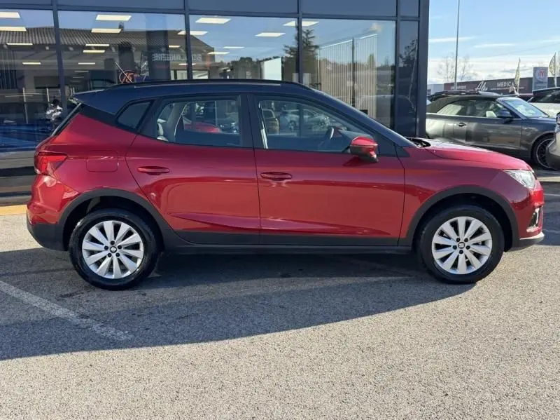 Vue de profil côté gauche d'un SEAT Arona rouge métal avec toit noir, garé devant une vitrine ensoleillée.