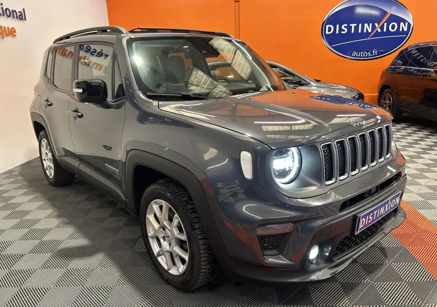 Jeep Renegade gris graphite en 3/4 avant droit, avec phares allumés et toit ouvrant visible en intérieur showroom.