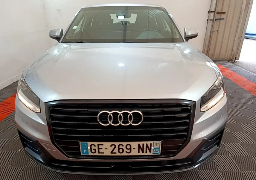 Vue avant d’un Audi Q2 gris clair 2020 avec calandre noire et phares LED allumés dans un garage.