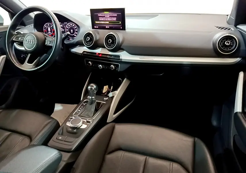 Intérieur Audi Q2 35 TDI 2020 vu côté conducteur, tableau de bord noir avec écran central et volant cuir multifonctions.