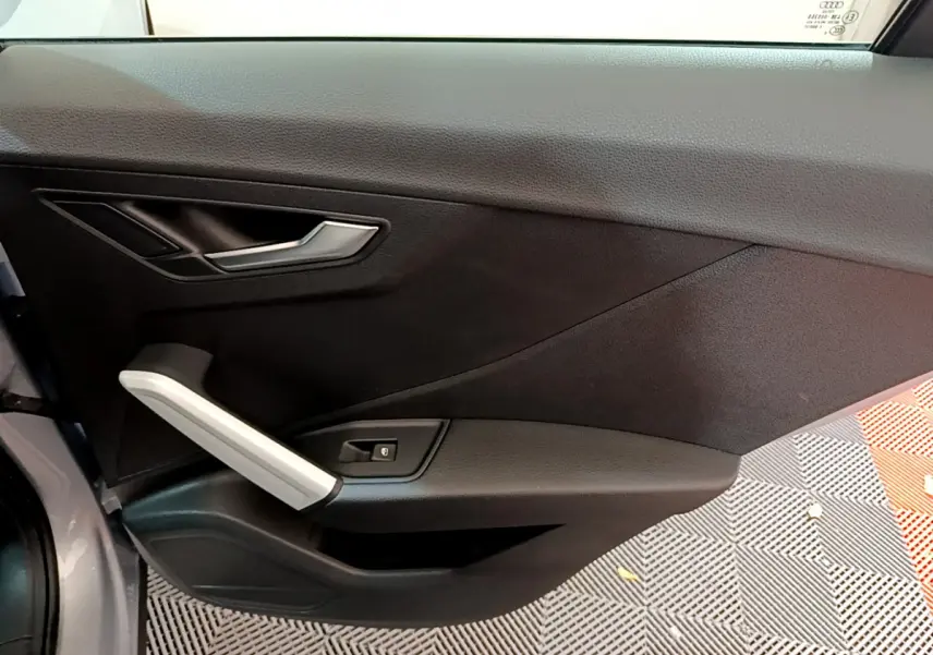 Vue intérieure du panneau de porte côté gauche de l'Audi Q2 35 TDI gris clair, avec poignée argentée et bouton de verrouillage.