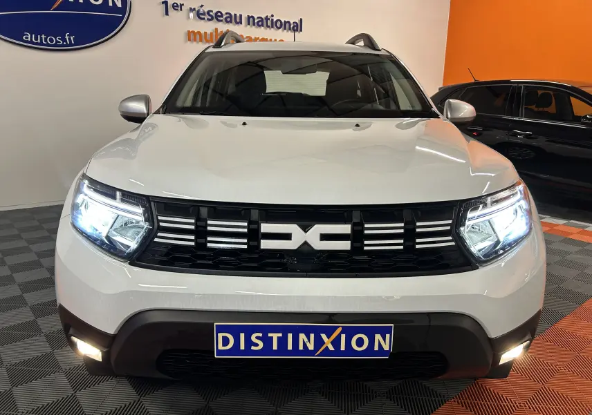 Vue avant d'un Dacia Duster blanc 2024 avec phares allumés et calandre noire distinctive en showroom.