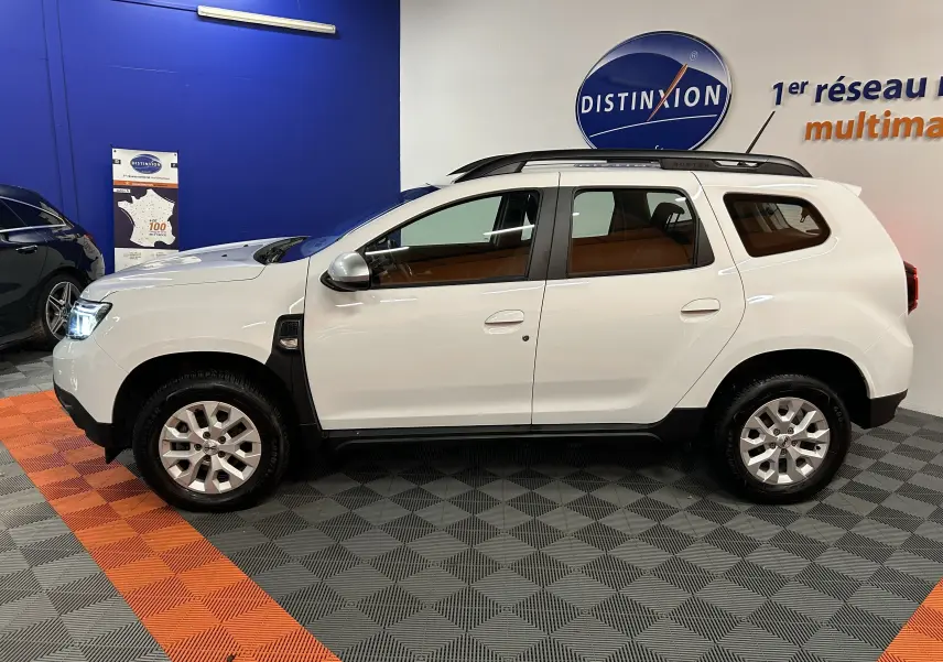 Vue de profil côté gauche d'un Dacia Duster blanc 2024 avec barres de toit et jantes alliage en intérieur showroom.