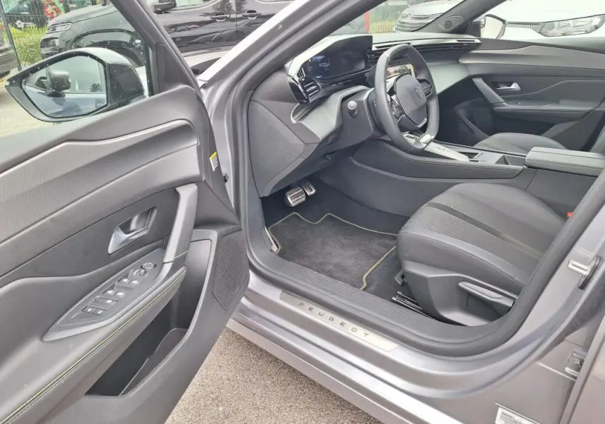 Vue intérieure côté conducteur d’une Peugeot 308 Hybrid 145 e-DCS6 GT gris, avec tableau de bord numérique et sellerie noire.