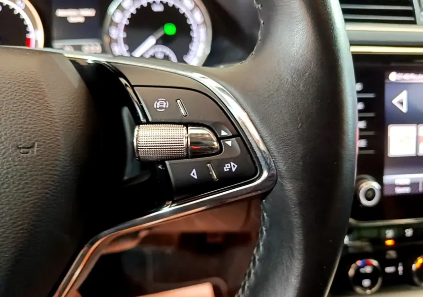 Gros plan sur les commandes gauche du volant cuir noir de la Skoda Superb 2021, avec tableau de bord et écran tactile en arrière-plan.