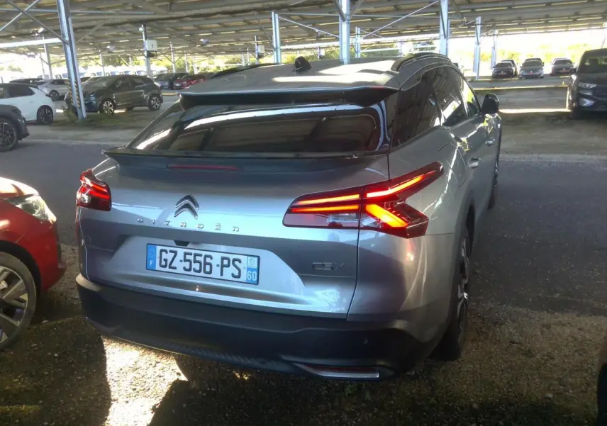 Vue 3/4 arrière d'une Citroën C5 X 2024 gris foncé avec feux arrière allumés, stationnée sous abri.