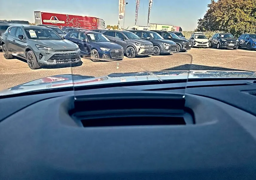 Vue depuis l'habitacle vers l'avant, tableau de bord et pare-brise, avec plusieurs voitures en arrière-plan sur un parking.