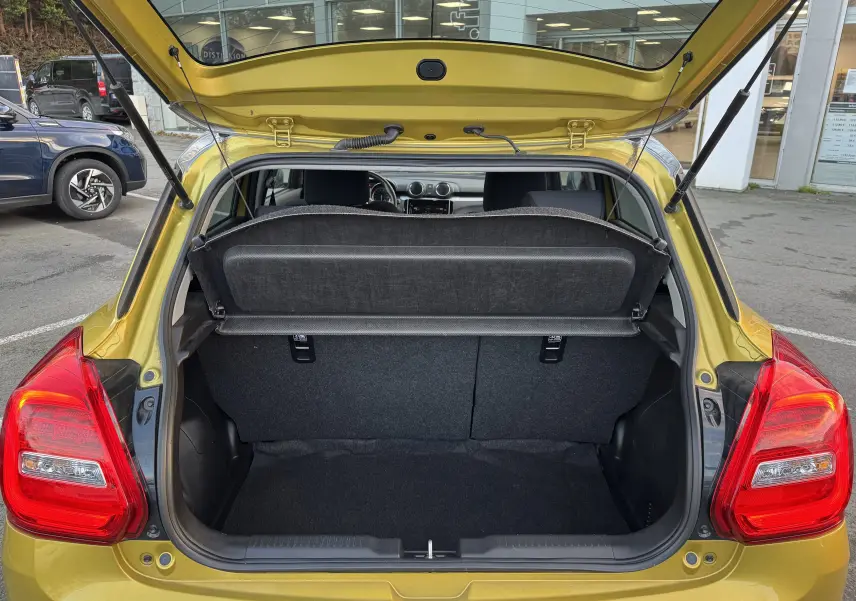 Coffre ouvert vu de l'arrière d'une Suzuki Swift 1.2 Pack Auto jaune/gris, montrant l'espace de chargement et l'intérieur arrière.