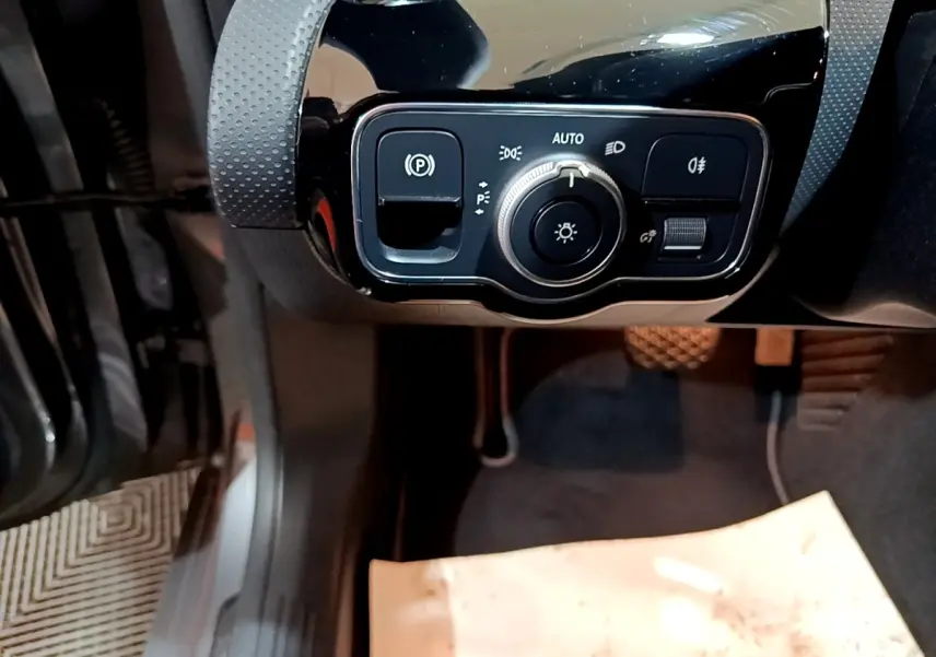 Gros plan sur les commandes d’éclairage et frein à main électrique côté gauche du tableau de bord noir brillant d’une Mercedes CLA 180 d gris clair.
