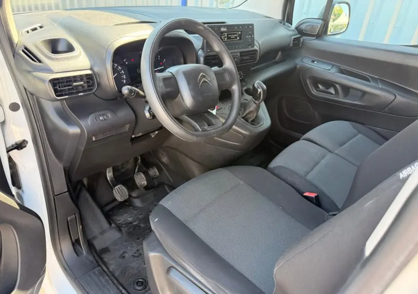 Vue intérieure côté conducteur du Citroën Berlingo blanc 2020, montrant le volant, tableau de bord et sièges tissu noirs.