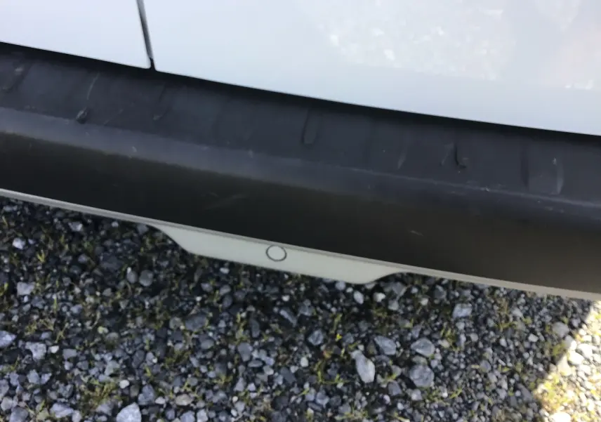 Gros plan sur le pare-chocs arrière blanc du Renault Kangoo 2024 avec capteur de recul visible sur fond de gravier.