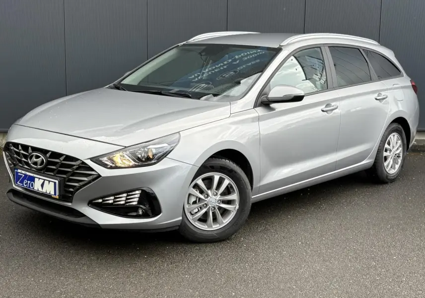 Hyundai i30 SW T-GDI 120 gris argent en 3/4 avant droit, avec jantes alliage et feux allumés.