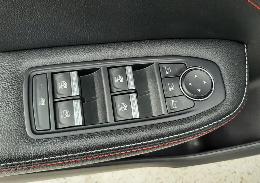 Gros plan sur les commandes des vitres électriques et des rétroviseurs sur la porte côté gauche, avec surpiqûres rouges sur cuir noir.