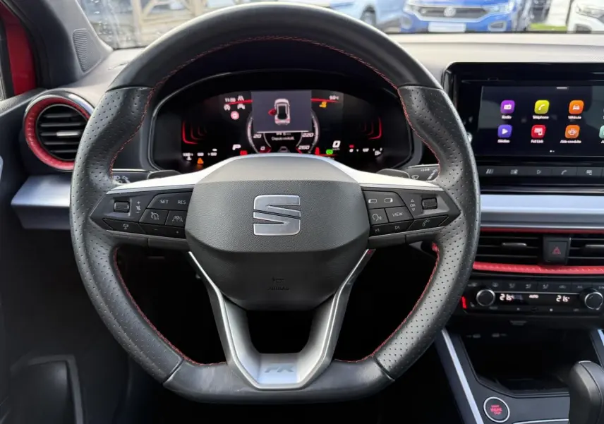 Vue rapprochée du volant sport en cuir perforé noir avec surpiqûres rouges et tableau de bord digital d'une SEAT Arona rouge 2024.