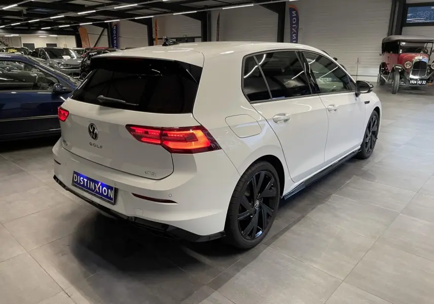 Vue 3/4 arrière droite d'une Volkswagen Golf 1.5 eTSI blanche avec jantes noires et vitres surteintées en intérieur showroom.