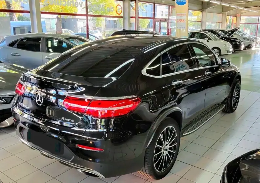Vue 3/4 arrière droite d’un Mercedes GLC Coupé 350 noir brillant avec jantes sport et feux arrière allumés en showroom.