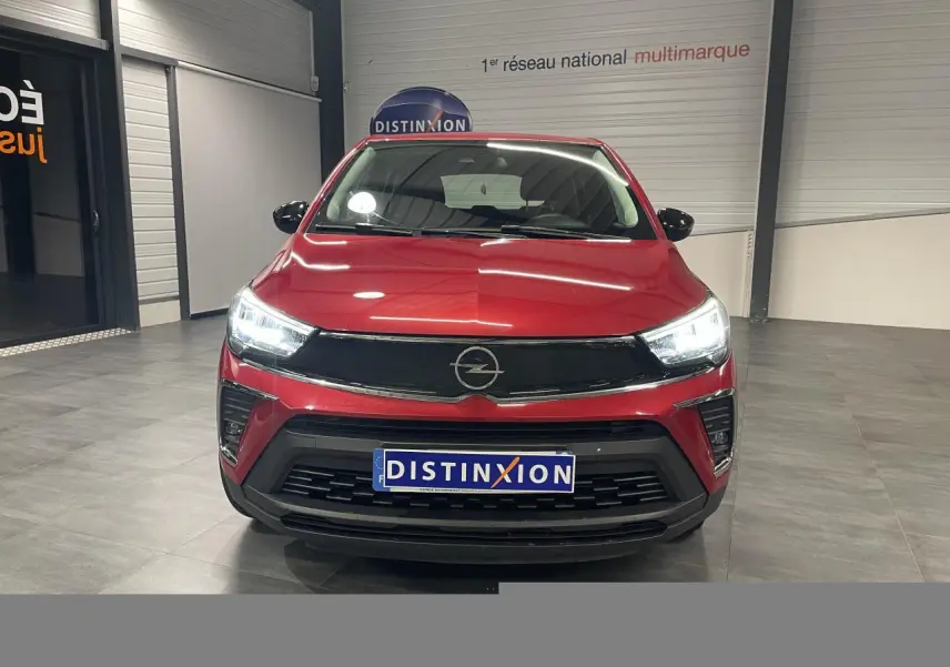 Vue de face de l’Opel Crossland rouge clair 2022 avec calandre noire et phares LED allumés en intérieur showroom.