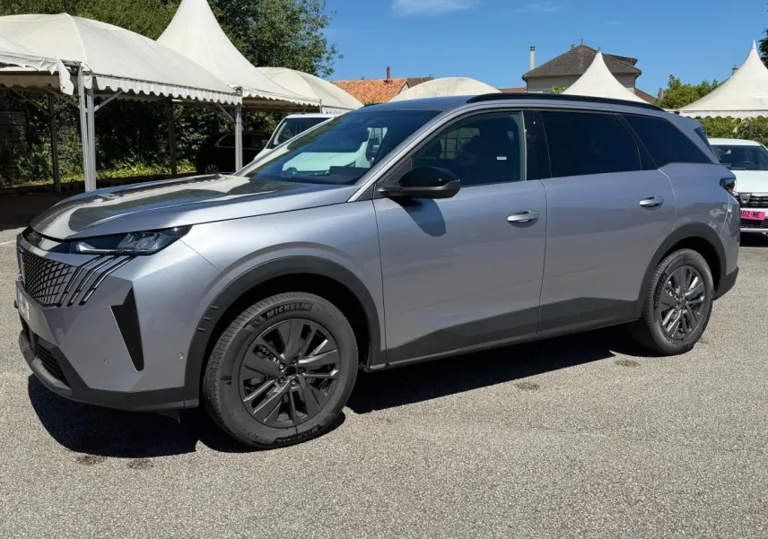 Peugeot 5008 gris clair vue 3/4 avant droit, avec calandre distinctive et jantes noires sur parking extérieur.