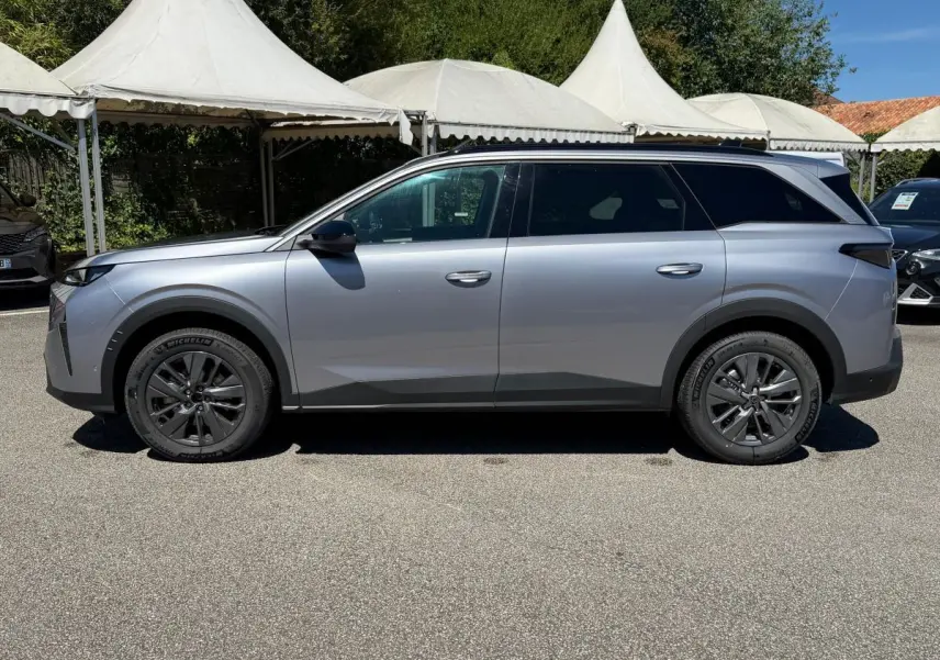 Vue de profil côté gauche d’un Peugeot 5008 gris clair 2025 avec jantes foncées et toit panoramique noir.
