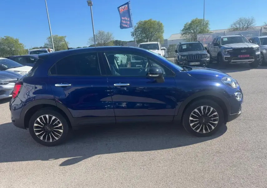 Profil côté gauche d'un FIAT 500X bleu MY23 avec jantes classiques 16 pouces, garé en extérieur par temps clair.