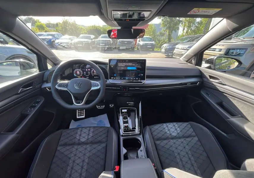Vue intérieure avant de la Volkswagen Golf E-TSI 150 DSG R-Line avec tableau de bord numérique et sièges tissu noirs.