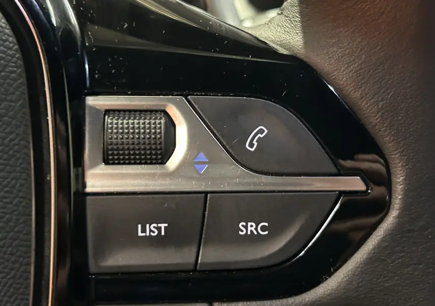 Gros plan sur les commandes noires du volant côté gauche du Peugeot 2008 gris Artense, avec boutons LIST, SRC et téléphone.