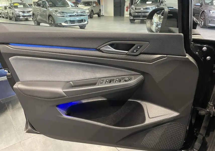 Vue intérieure côté gauche de la porte d’une Volkswagen Golf 2024 noire avec éclairage d’ambiance bleu et commandes électriques.