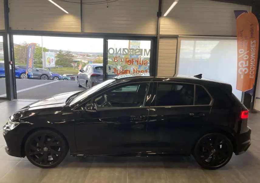 Profil côté gauche d'une Volkswagen Golf 8 R-Line noire métallisée avec jantes Bergamo noires en intérieur showroom.