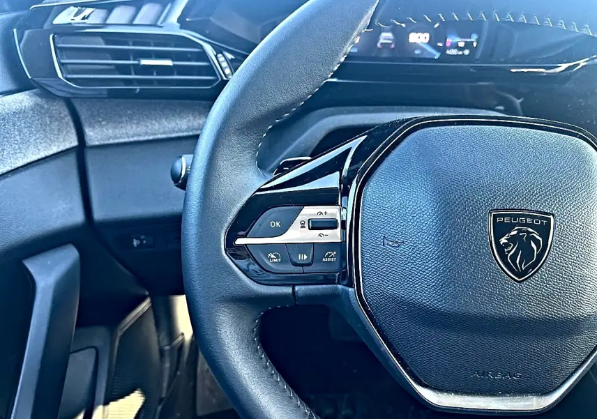 Vue rapprochée du volant multifonction noir de la Peugeot 308 2025 avec tableau de bord numérique en arrière-plan.