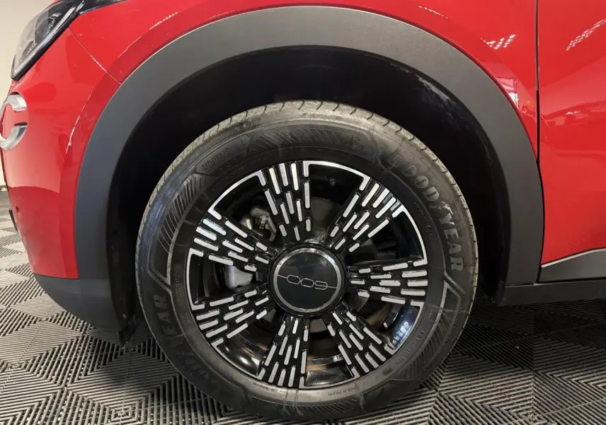 Gros plan sur la roue avant gauche d’une FIAT 600 rouge foncé avec jante noire et motifs argentés.