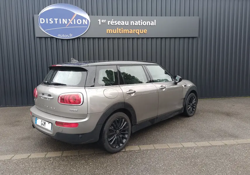 Vue 3/4 arrière droite d'une MINI Clubman F54 Silver Metal avec toit noir et jantes noires, stationnée devant un mur gris.