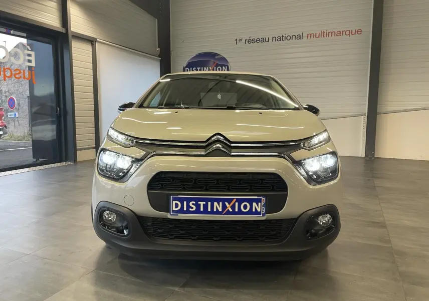 Vue avant d'une Citroën C3 beige clair 2024 avec feux LED allumés et calandre noire en intérieur showroom.