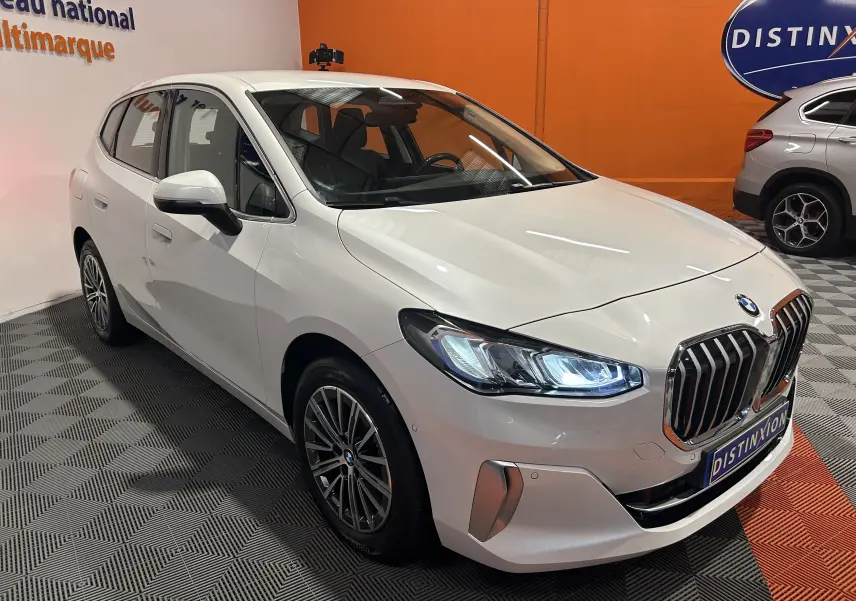 BMW Série 2 Active Tourer blanc vue 3/4 avant droit avec phares LED allumés dans un showroom.