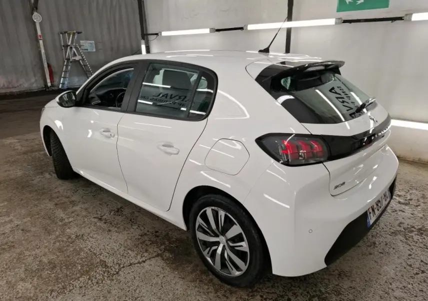 Peugeot 208 blanche vue 3/4 arrière droit en intérieur, avec jantes alliage et feux arrière distinctifs noirs.
