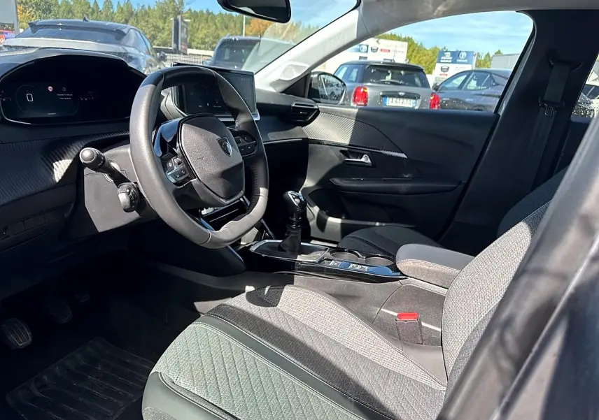 Intérieur avant droit de la Peugeot 208 1.2 100 Allure 2025, volant compact et sièges tissu gris, avec levier de vitesse manuel.